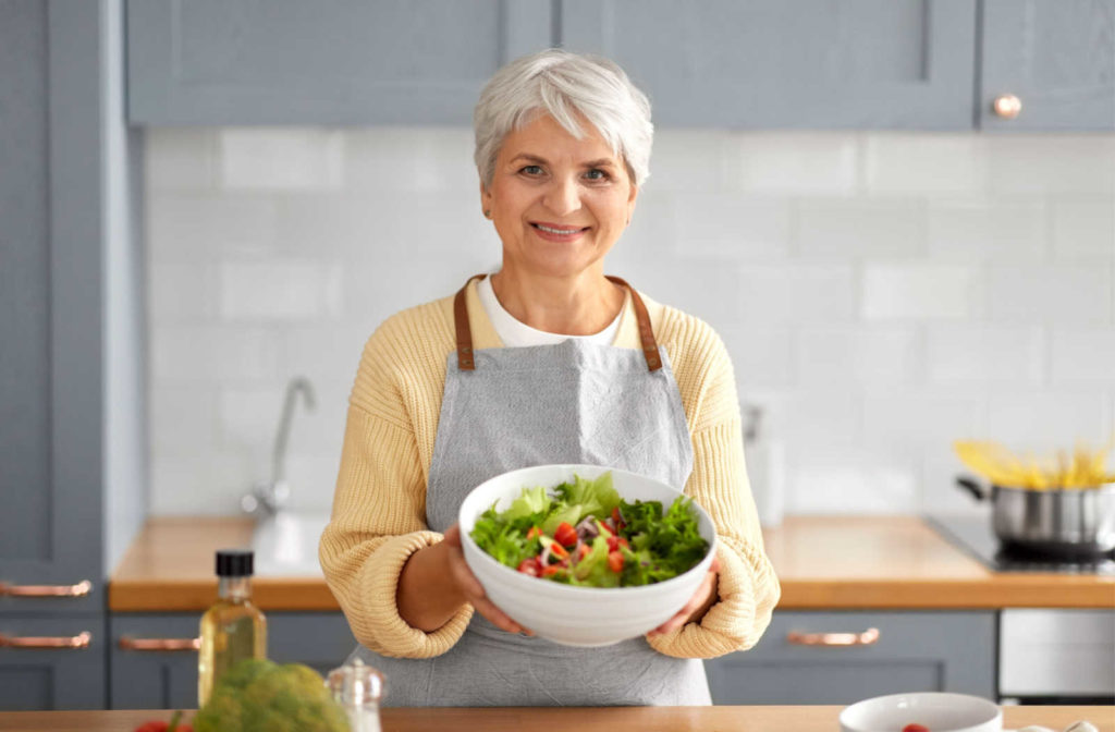 A senior woman smiling and holding a bowl of salad in the kitchen.