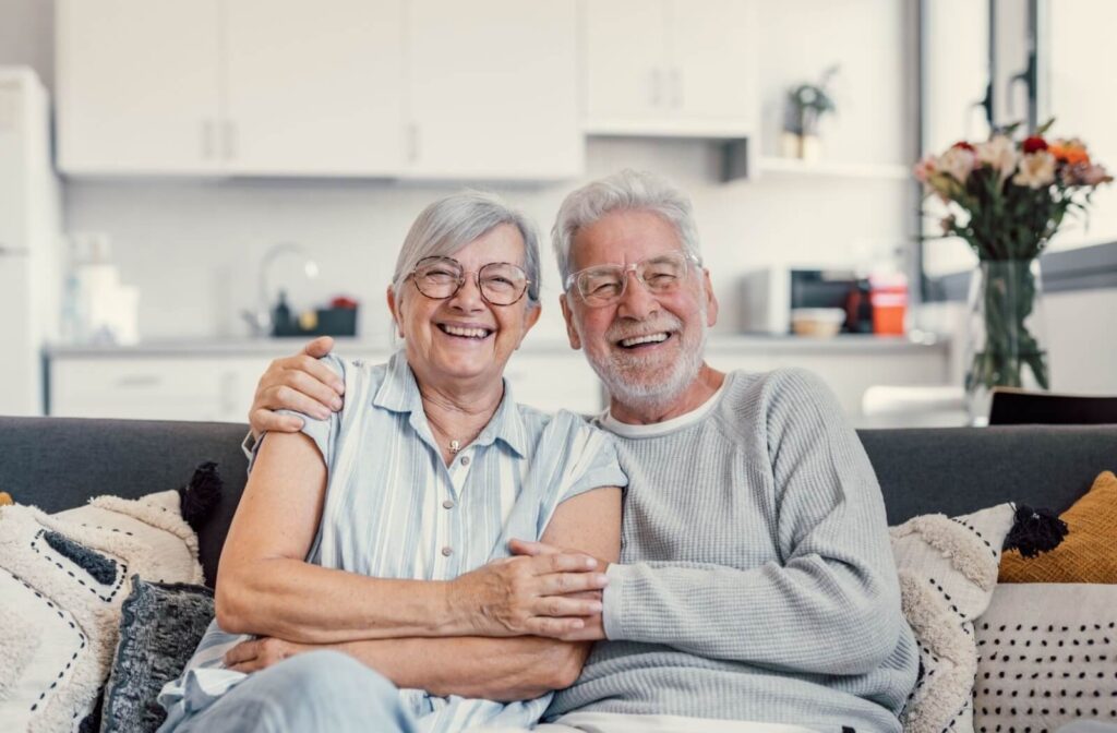 An older couple smiles with arms around one another while sitting on the couch in their new assisted living apartment