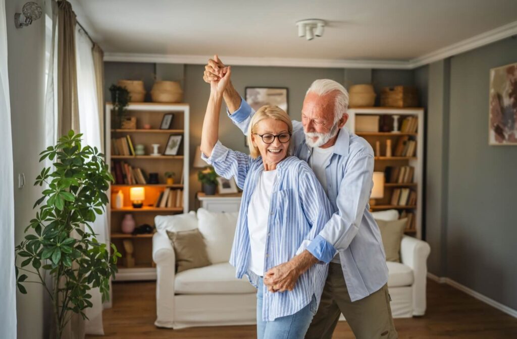 An older couple laughs and dances together in their new well-decorated living room in assisted living